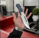 In a modern kitchen, a person with a watch holds the Texan Knives Premium D2 Steel Hunting Knife, featuring a blue and gold patterned handle. A potted plant, stove, and the branded leather case from Texan Knives are visible in the background.