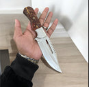 A hand holds the Texan Knives D2 Steel Hunting Knife with a pine cone handle and silver blade, over a light wooden surface.