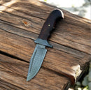 The Texan Knives Premium Damascus Steel Hunting Knife with Rosewood Handle (9” overall) rests on a weathered wooden surface in sunlight, highlighting its expert craftsmanship and making it an essential hunting tool.