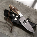 The Texan Knives Texas Knives Premium Damascus Steel Hunting Knife with Stag Horn Handle (10" overall) rests on a black leather sheath with a braided brown lanyard, displayed on textured gray stone—ideal for outdoor adventures.