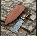 The Texan Knives Premium Damascus Steel Hunting Knife features a stag horn handle and engraved blade, displayed on stone alongside its ornate brown leather case.