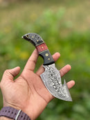 A hand holds the Texan Knives Premium Custom Handmade Damascus Steel Guthook Skinning Knife, featuring a 7.5” sharp blade with wavy designs and a resin handle, against a blurred green foliage background. Leather sheath included.