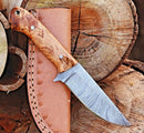 The Texan Knives Premium 10-Inch Damascus Steel Hunting Knife with a rosewood handle rests on its leather sheath atop cut logs, making it an ideal gift for any outdoorsman.