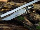 The Texan Knives 13-Inch Damascus Steel Cowboy Bowie Knife with a black and silver buffalo horn handle rests on weathered wood by a metal ruler, displaying its full blade length. The scene includes green moss for a rustic touch.