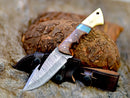 The Texan Knives Premium Damascus Guthook Skinning Knife features a 10" patterned blade and a rosewood & bone handle, resting on a leather sheath atop animal fur with rough bark in the background.