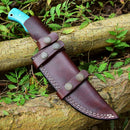 The Texan Knives Custom Handmade Damascus Steel Hunting Knife, featuring a 10-inch blade and resin handle, sits in its leather sheath adorned with metal studs on a tree branch among green leaves.