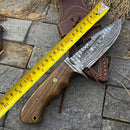 The Texan Knives 🔥 Handmade Damascus Steel Hunting Knife – Rosewood Handle & Damascus Guard + Leather Sheath, about 23 cm long, rests beside its sheath on stone tiles with a yellow measuring tape and dried grass in the background.