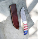 The Texan Knives Premium Guthook Skinning Knife features a 9" Damascus steel blade, a Texas flag handle, and comes with a brown leather sheath, displayed on a light stone surface.