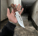 A hand holds the Texan Knives Premium J2 Steel Hunting Knife with Pain Cone Handle and Leather Belt Loop Case, its gleaming blade contrasting against a weathered stone wall—showcasing Texan Knives’ top-tier hunting gear.