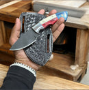 A hand holds the Texan Knives Premium J2 Steel Hunting Knife (6.25") featuring a blue, white, and red Texas flag handle and a wide blade, paired with a textured black leather case, set against a wooden background.