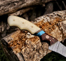 A close-up of the Texan Knives Fishing Fillet & Boning Knife – 13" Damascus steel blade with a camel bone and rosewood handle, shown outdoors on a log, includes blue accents and comes with a leather sheath.