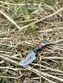 The Texan Knives Premium Custom Handmade Damascus Steel Guthook Skinning Knife with a resin handle and 7.5” sharp blade rests outdoors on dry grass, highlighting its patterned blade and leather sheath.