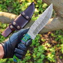 A gloved hand holds the Texan Knives Custom Handmade Damascus Skinning Knife – 4.5" Resin Handle Blade for Seasonal Hunts with a green and blue handle; a brown leather sheath sits on a tree branch, foliage below.