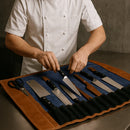 A chef in a white double-breasted coat arranges knives in the Aaron Leather Goods Leather Knife Roll on a metal surface, keeping kitchen knives and scissors organized and protected for busy kitchen environments.