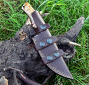The Custom Handmade Damascus Steel Skinner Knife with Sheep Horn Handle – 4.5" Blade by Texan Knives rests in a brown leather sheath with three snap-button straps, displayed on a tree stump amidst green grass.