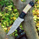 The Texan Knives Custom Handmade Skinning Knife Set features a 4.5" patterned Damascus blade and a unique camel bone & resin handle, showcased resting on a branch with softly blurred green foliage in the background.