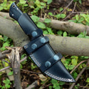 The Texan Knives Custom Handmade Skinning Knife, featuring a Damascus steel blade and a buffalo horn & camel bone handle, sits in its leather sheath on a tree branch, surrounded by green foliage and twigs.