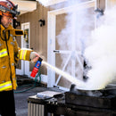 A firefighter in uniform uses the Prepared Hero Fire Spray by Prepared Hero to extinguish thick smoke from a barbecue grill outside a house, demonstrating key kitchen fire safety practices.