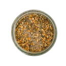 A glass jar of SALTOPIA Infused Sea Salts' PRESTO PESTO Basil, Garlic & Lemon Salt sits on a white background, showcasing its coarse mix of dried citrus peel pieces and salt when viewed from above.