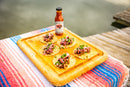 Six tacos topped with meat, onions, and cilantro are arranged on a wooden tray next to a bottle of Brushfire Farms Prickly Pear BBQ Sauce on a colorful striped cloth, outdoors by a body of water.