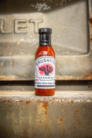 A bottle of Brushfire Farms Prickly Pear BBQ Sauce from Brushfire Farms rests on a rusty metal surface, with a blurred metal plate visible in the background.
