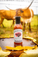 A bottle of Brushfire Farms Prickly Pear BBQ Sauce from Brushfire Farms rests on a rusty surface alongside cactus peels, as two cows graze behind a wire fence in the blurred, grassy background.