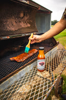A person brushes savory BBQ sauce onto ribs cooking on a grill. A bottle of Brushfire Farms Prickly Pear BBQ Sauce sits on the metal grill shelf. A rustic shed and grass are visible in the background.