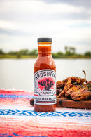 A bottle of Brushfire Farms Prickly Pear BBQ Sauce from Brushfire Farms rests on a vibrant outdoor rug, accompanied by grilled meat with a scenic lake in the background.