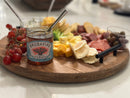 A wooden board with sliced cheeses, crackers, meats, grapes, olives, and an open jar of Brushfire Farms Prickly Pear Pepper Jam with a spoon, set on a light countertop.