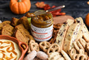 A jar of Terrapin Ridge Farms Pumpkin Butter is surrounded by biscotti, sliced pumpkin bread, pretzels, cookies, and caramel-swirled dip, with mini pumpkins and leaves creating a festive fall entertaining display.