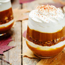 A glass dessert layered with pumpkin puree, crumbled nuts, and whipped cream is topped with SALTOPIA’s Pumpkin Cappuccino Infused Sugar: PUMPKIN TO TALK ABOUT. The treat rests on a wooden table adorned with autumn leaves.