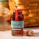 A glass jar of Honey Truck Co Raw Florida Wildflower Honey with a red lid sits on a table next to a wooden honey dipper. In the background, a wicker basket and another glass container are softly blurred. Sunlight illuminates the scene, bringing out the golden hues of this local wildflower honey from Honey Truck Co.