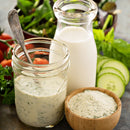 A wooden bowl of SALTOPIA Infused Sea Salts' Ranch Seasoning Low Sodium: SHOOK UP sits near a jar of creamy ranch, a glass bottle of milk, and fresh cucumber, tomato, and greens in the background.