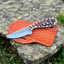 The Texan Knives Ranch Cowboy Knife features a patterned blade and textured sheep horn handle, resting on its orange leather case atop rough stone outdoors—showcasing the rugged style of Texan Knives.