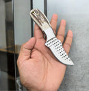 A person holds the Texan Knives Texas Knives Ranger’s Legacy Cowboy Knife with a stag horn handle and patterned blade featuring black triangular markings, indoors near a glass surface.