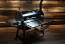 Black pellet grill with a Pimp My Grill® Tactical Stainless Folding Front Shelf attached for extra prep space.