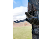 A person in camouflage clothing and gloves holds a DALSTRONG Fishing Hook Knife 5" from the Night Shark Series (NSF Certified) outdoors, with blurred grass and sky visible in the background.