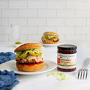 A chicken sandwich with lettuce, cheese, and pickles sits on a plate next to a jar of Terrapin Ridge Farms Red Pepper Jelly. Another sandwich is blurred in the background on a white tile kitchen counter.