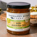 A jar of Terrapin Ridge Farms Pear Pistachio Jam rests on the counter, its label showing it's gluten free, vegan, and made with simple ingredients. A bread slice—ideal for charcuterie boards—appears in the background.