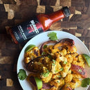 A plate of roasted cauliflower, red onion, lime wedges, and cilantro sits beside a bottle of Bravado Spice Ancho Masala Scorpion-Reaper Hot Sauce—an Indian-spiced hot sauce from Bravado Spice—on a wooden surface.