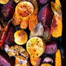 Colorful roasted carrots, beets, red onion, garlic, sliced lemon, yellow tomatoes, and bay leaves on a baking tray sprinkled with SALTOPIA Infused Sea Salts' ORANGE YOU TASTY Orange & Garlic Salt for a vibrant citrus flavor burst.