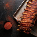 A rack of seasoned, cooked ribs on a metal tray sits on a dark surface with a cup of Saltopia Habañero Salt: HOT HEAD by SALTOPIA Infused Sea Salts and scattered spice nearby.
