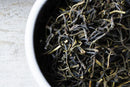 Close-up of World Spice Rwandan Green loose-leaf tea in a white bowl, placed on a light-colored surface. The dark, twisted leaves are mountain-grown green tea from Rwanda, highlighting the unique qualities of World Spice’s Rwandan Green.