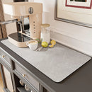 A beige espresso machine, a glass pitcher, lemons, a knife, and a cappuccino with latte art sit on the Lovely Home Icon Coffee Machine Mat—a rubber backing, water-absorbent dish drying pad—atop a dark cabinet, adding a touch of modern table decoration to the gray countertop.