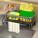 A hand places a scrub brush into the Lovely Home Icon Multifunctional Sponge Draining Rack – Kitchen Cloth & Dishwashing Liquid Storage, which holds a sponge, scrub pad, large bottle of cleaner, and has a striped towel hanging in front. The organizer sits on the kitchen countertop above the sink.