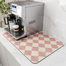 A silver espresso machine sits on a counter atop the Lovely Home Icon Coffee Machine Mat, a water-absorbent dish drying pad featuring rubber backing and a pink and cream diamond pattern that adds modern flair to the kitchen. A coffee cup with layered froth rests on the drip tray, while a small plant is visible in the background.