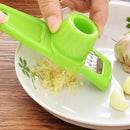 A hand uses the Lovely Home Icon 1PCS Garlic Crusher Press Multi-Functional Manual Ginger Garlic Grinding Grater Cutter and Peeler to grate garlic over a white plate, with garlic cloves and a bulb placed nearby.