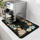 A silver espresso machine sits on a white counter with a cup of cappuccino. Underneath is the Lovely Home Icon Coffee Machine Mat, a water-absorbent dish drying pad with rubber backing, decorated in colorful floral designs that add a modern touch to the space. A green plant accents the corner beside the marble-patterned backsplash.