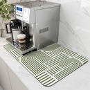 A silver espresso machine sits on a white countertop next to a plant, with a cup of coffee topped with milk foam resting on the drip tray. Underneath the machine, the Lovely Home Icon Coffee Machine Mat—a green and white geometric-patterned, water-absorbent dish drying pad with rubber backing—adds a modern decorative touch to the kitchen worktop.