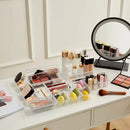 A vanity table showcases the Lovely Home Icon 10-Piece Versatile Drawer Organizer Set, featuring clear plastic trays and dividers in four sizes to perfectly store makeup brushes, lipsticks, bottles, sponges, and palettes. A round mirror and various cosmetics are neatly arranged on the white tabletop, with the organizer set keeping everything in flawless order.